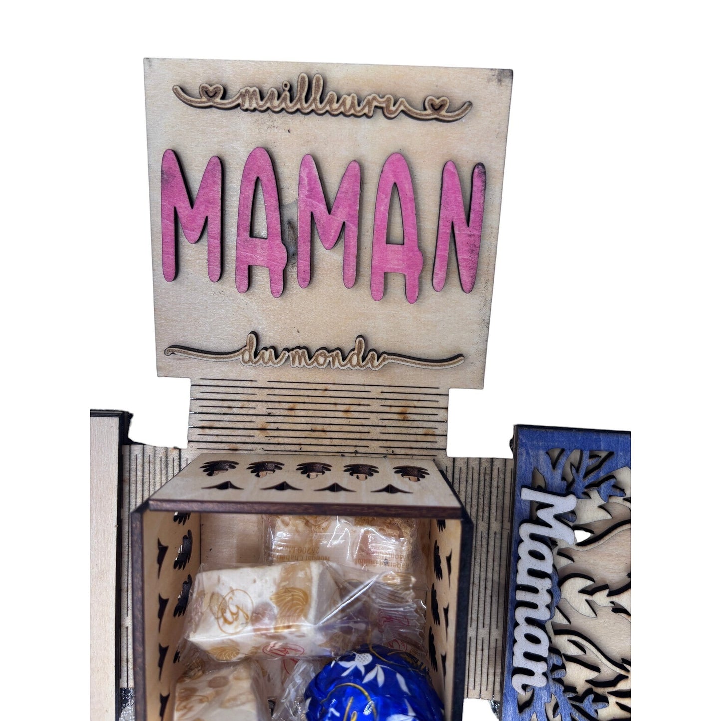 Boîte cadeau surprise en bois L'Atelier de Lylou pour la fête des mères, avec "Meilleure Maman du monde" gravé et des compartiments remplis de nougat et d'une boule bleue.