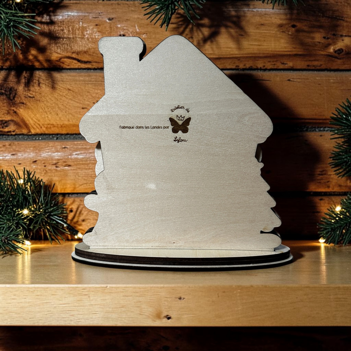 Dos de la maison de Noël en bois L'Atelier de Lylou avec gravure artisanale sur fond de branches de sapin