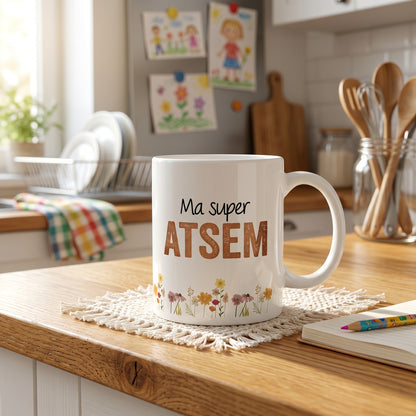 Mug blanc L'Atelier de Lylou avec l'inscription "Ma super ATSEM" et un motif floral coloré. Cadeau personnalisé pour ATSEM.