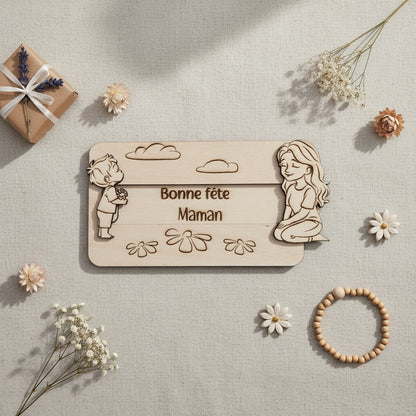 Plaque en bois L'Atelier de Lylou "Bonne fête Maman" avec un enfant offrant des fleurs et une femme assise, décorée de nuages et de fleurs.