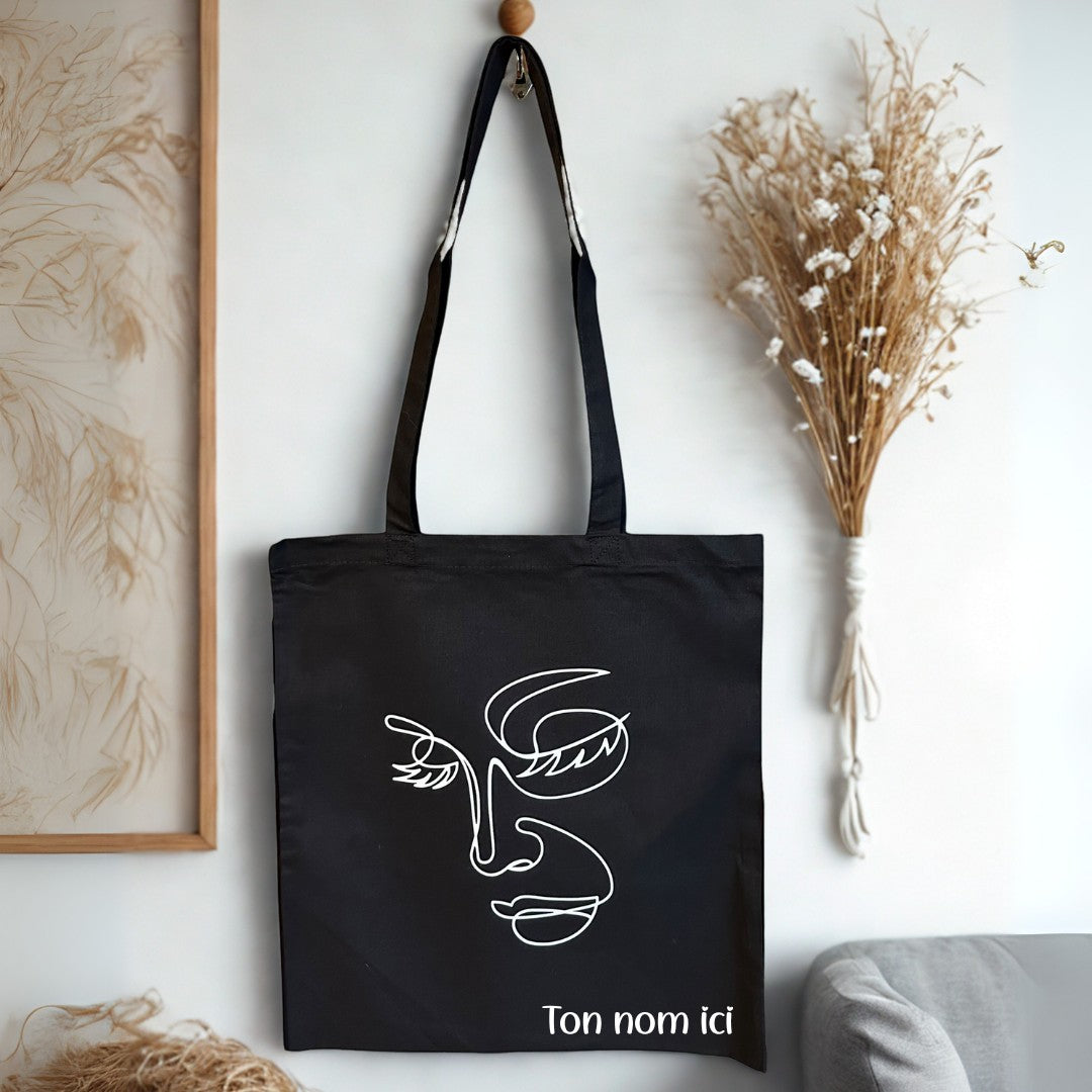 L'Atelier de Lylou tote bag noir en coton avec motif visage abstrait et personnalisable.