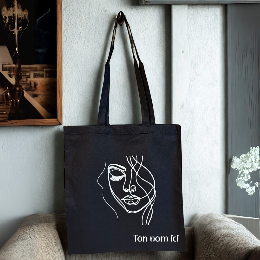 Tote bag noir en coton L'Atelier de Lylou avec un dessin de visage abstrait et personnalisable avec votre nom.